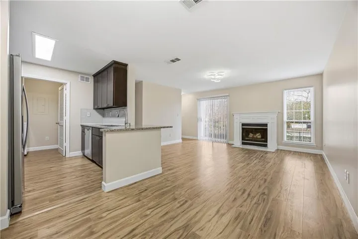 Open-concept living area featuring wood-finish flooring throughout, a kitchen with dark cabinetry and stainless steel appliances, a centered island with prep space, and a fireplace with surround shelving