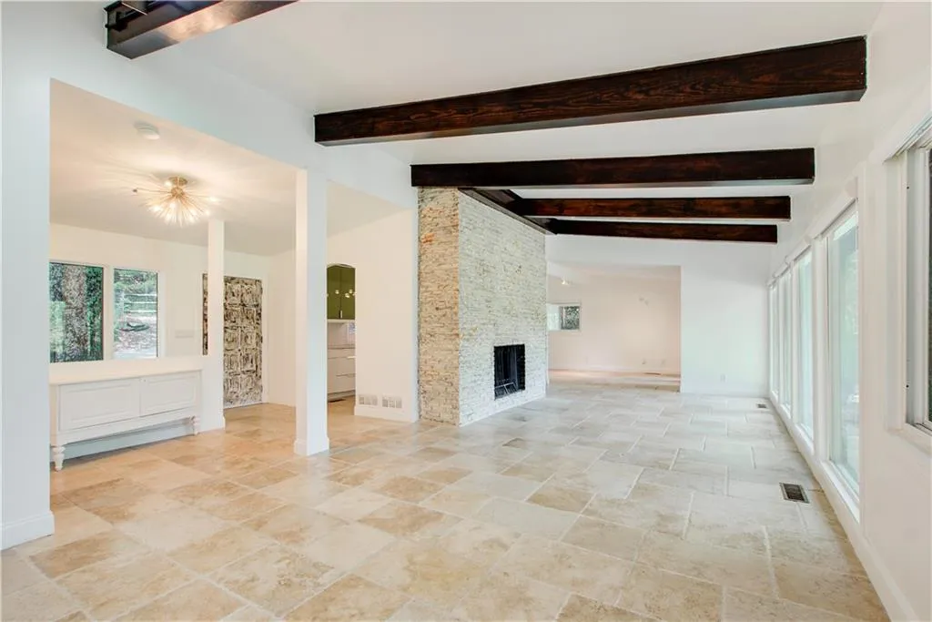 Unfurnished living room featuring stone tile floors, baseboard heating, baseboards, beam ceiling, and a stone fireplace