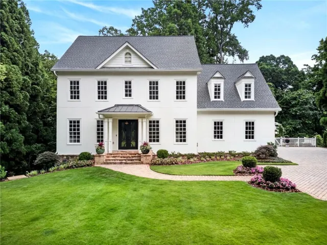 Traditional architecture featuring a white exterior, dark shingle roof, symmetrical window placement, and a brick paver driveway