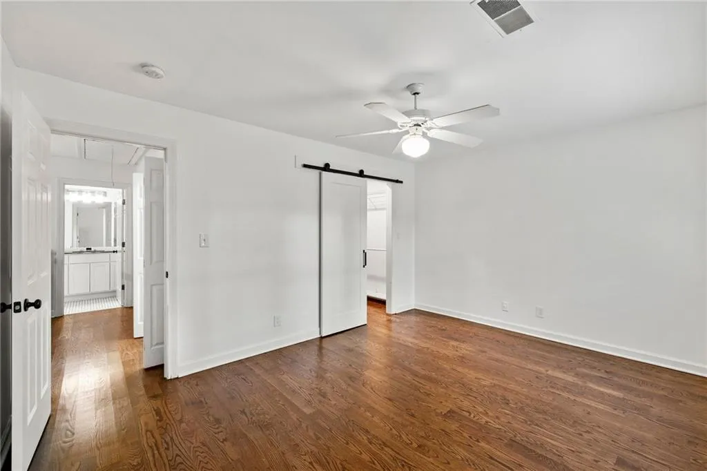 Unfurnished bedroom with a barn door, dark wood-style floors, ensuite bathroom, and a ceiling fan