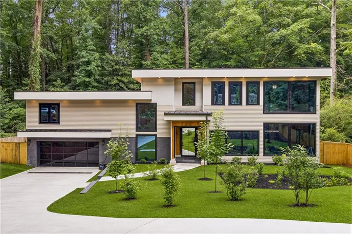 Modern architectural design featuring expansive dark-framed windows, a sleek garage door, a concrete driveway, and a manicured lawn with mature landscaping