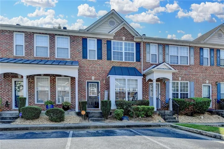 Brick facade townhome featuring a covered porch entrance, bay window, white window frames with dark blue shutters, and mature front landscaping