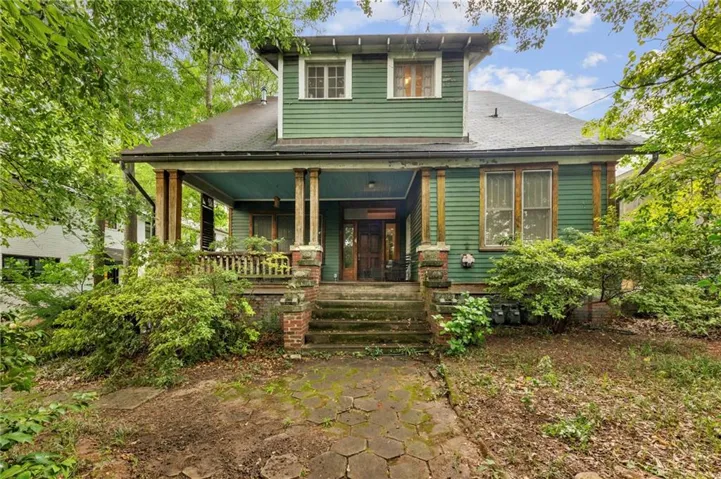 Green siding home featuring a covered front porch with brick and wood columns, a hexagonal paver walkway, and mature perimeter landscaping