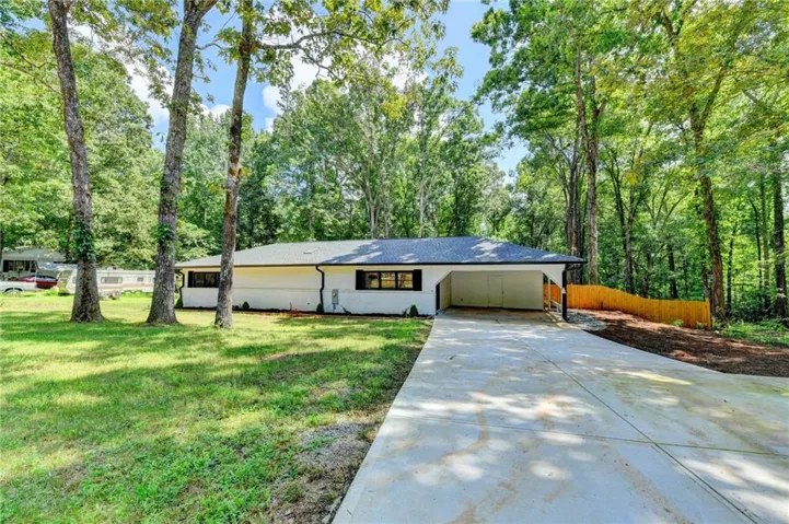 White exterior with black trim, concrete driveway, attached carport, wood privacy fence, and mature trees