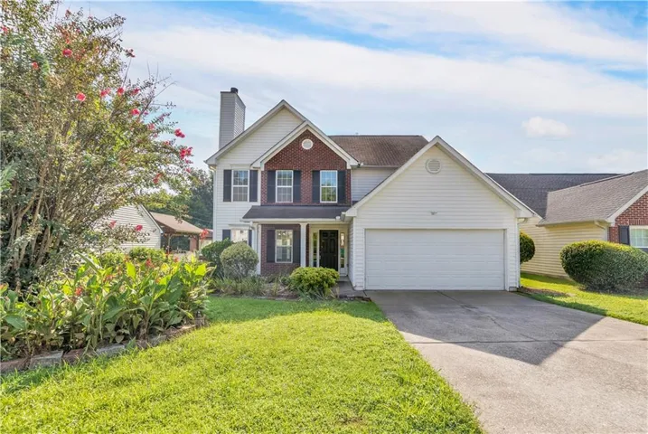 Two-story Colonial-style residence featuring a brick and siding exterior, two-car attached garage with white overhead doors, front-facing gable roofline, and a manicured front lawn bordered by mature landscaping and flowering shrubs