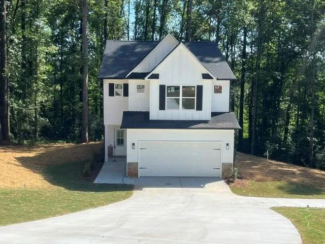 White vertical siding with black shutters, attached two-car garage with stone accents, black shingle roof, concrete driveway, and mature trees surrounding the property