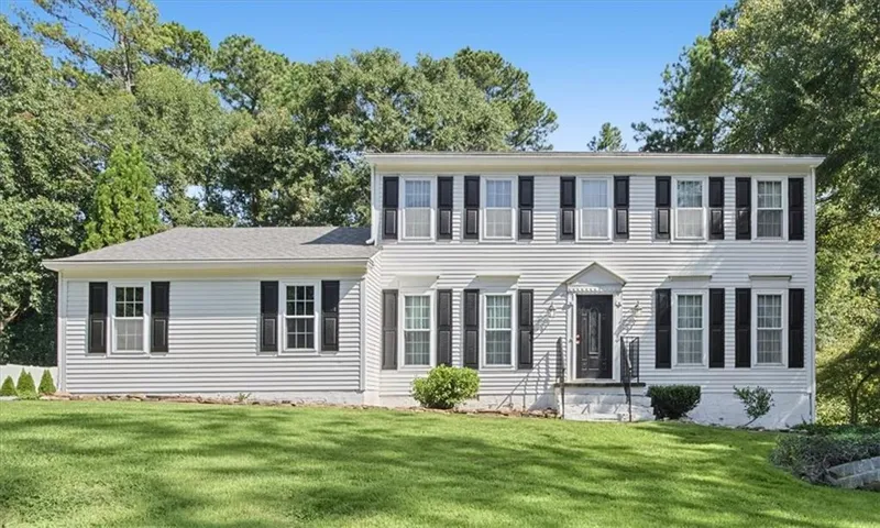 White siding exterior with a dark entry door, multiple windows, and black shutters, set against a backdrop of mature trees and a manicured lawn
