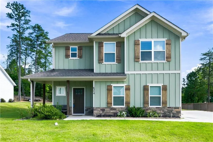 Two-story residence featuring green board-and-batten siding, stone facade accents, wood shutters, a covered front porch, and a dark entry door