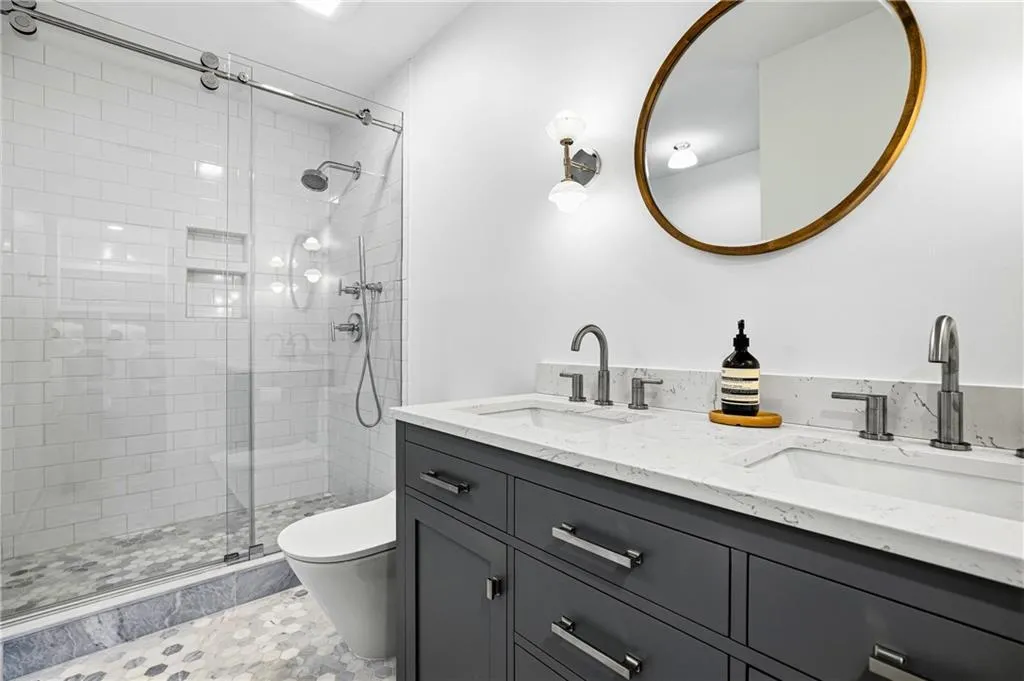 Contemporary bathroom featuring a dual vanity with a white countertop and undermount sinks, a frameless glass shower enclosure with subway tile surround, hexagonal floor tile, and a wall-mounted oval mirror