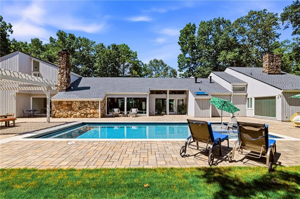 Outdoor swimming pool with integrated spa and paver surround, complemented by a stone-clad exterior, multiple large windows, and a pergola-covered patio area