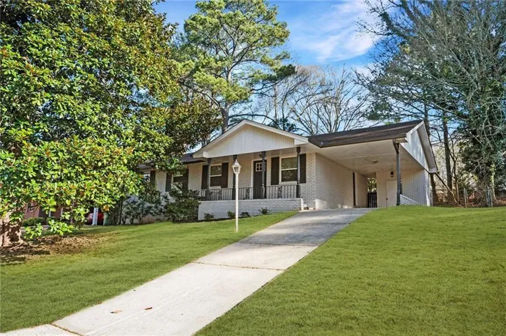 Exterior featuring a light brick facade, a portico entry with black railing, a paved driveway, a carport, and mature trees
