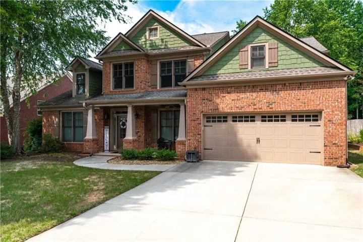 Brick exterior with green shake siding accents, a two-car garage, covered porch with columns, a curved concrete walkway, and mature landscaping