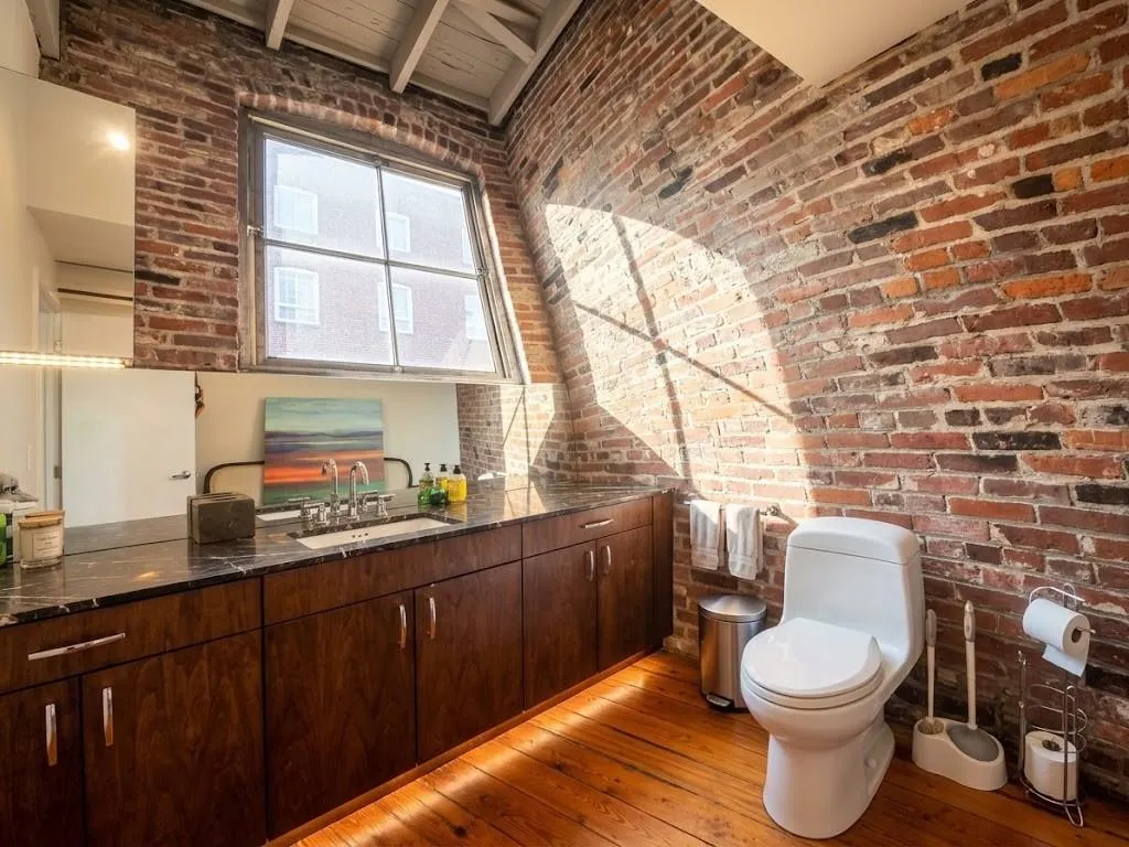 Exposed brick wall, natural wood flooring, wood-finish cabinetry, built-in vanity with integrated sink, and overhead window