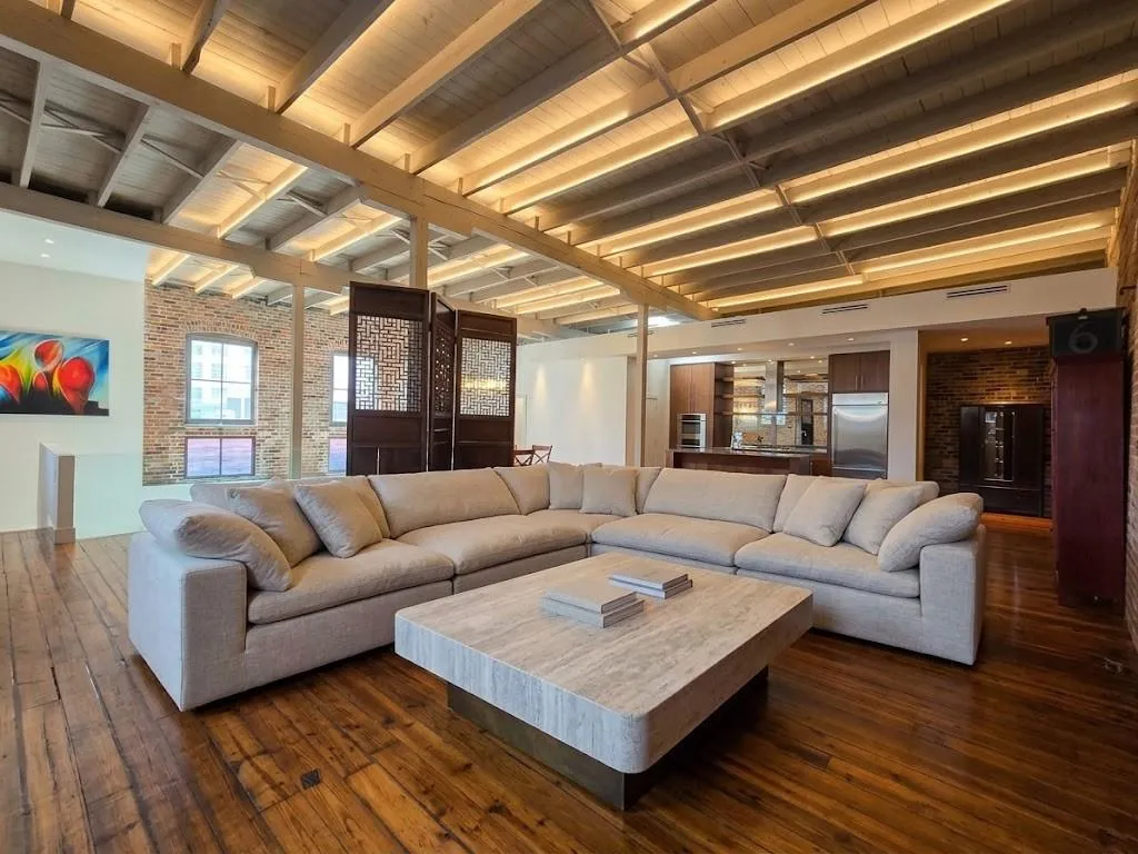 Exposed wood beam ceiling with integrated lighting, wood-finish flooring, exposed brick wall, built-in kitchen cabinetry with stainless steel appliances, virtual staging living room furniture and LED lighting