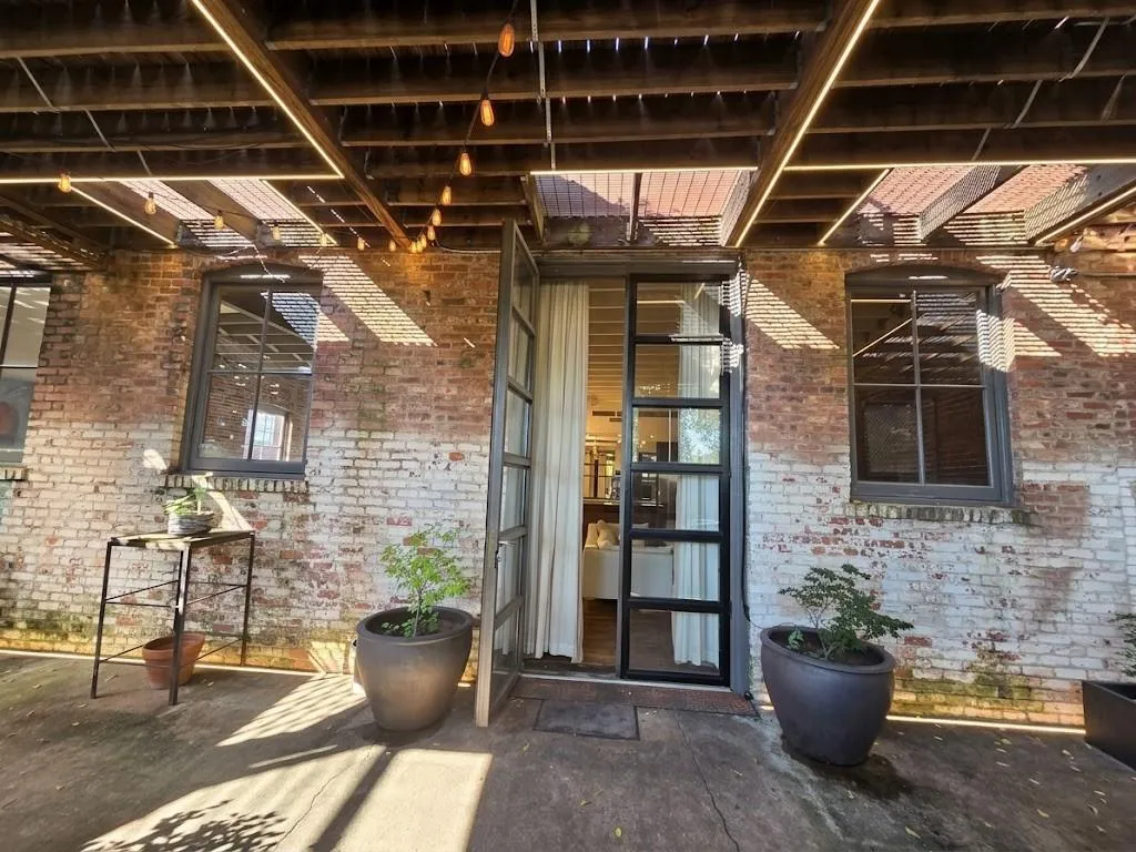 Exposed brick wall exterior, open-beam wood ceiling, industrial-style glass entry door, string lighting, and concrete patio flooring