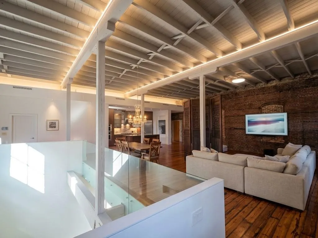 Exposed brick wall, hardwood flooring, open ceiling with exposed beams, glass panel railing, and built-in kitchen cabinetry, virtual LED lighting, sofa and Wall TV