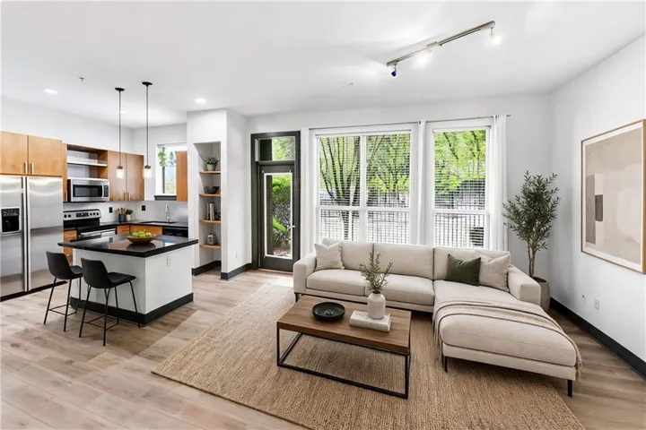 Open-concept living space featuring wood-finish flooring, a kitchen island with pendant lighting, stainless steel appliances, and large windows with blinds