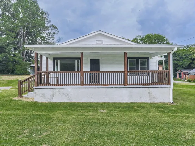 White siding exterior, covered front porch with wood railings, dark-framed windows, dark entry door, and a grass lawn