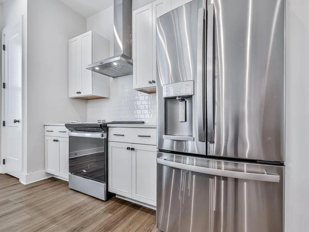 Kitchen featuring appliances with stainless steel finishes, wall chimney exhaust hood, light wood finished floors, decorative backsplash, and white cabinetry