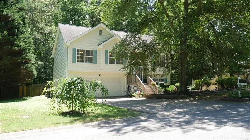 Split-level residence featuring light-colored siding, contrasting shutters, an attached two-car garage, an elevated entry with wood stairs, and mature trees providing shade