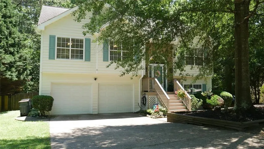 Split-level residence featuring light-colored siding, contrasting shutters, an attached two-car garage, an elevated entry with wood stairs, and mature trees providing shade