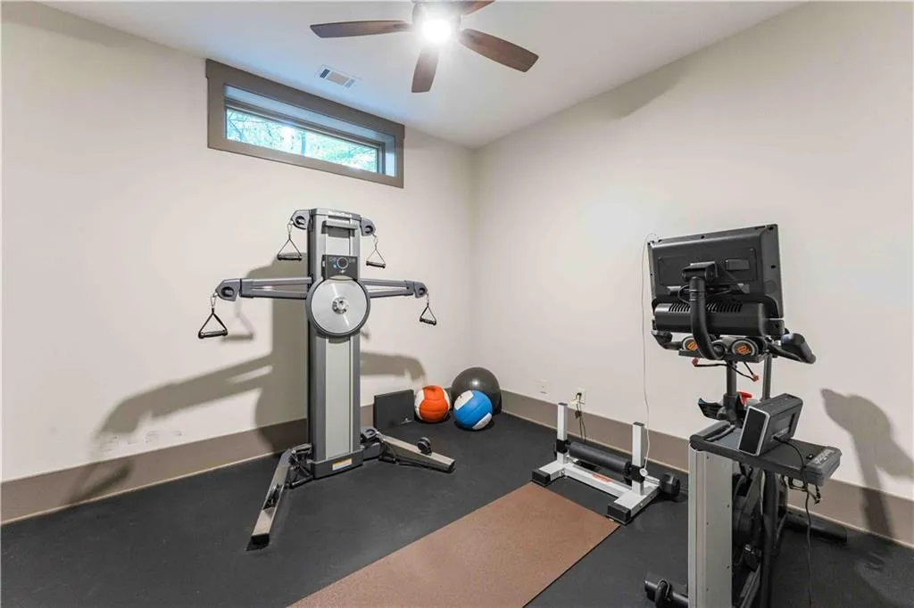 Dedicated exercise room featuring dark rubber flooring, a horizontal window with dark trim, a ceiling fan with integrated lighting, and neutral-toned walls