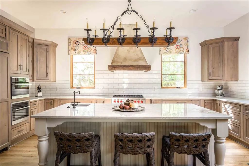 Expansive kitchen featuring light wood-finish cabinetry, a large center island with a light-toned countertop, a range with a decorative hood, built-in double ovens, and a white subway tile backsplash