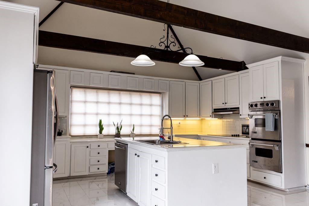 Kitchen featuring appliances with stainless steel finishes, light tile floors, beam ceiling, white cabinetry, and an island with sink