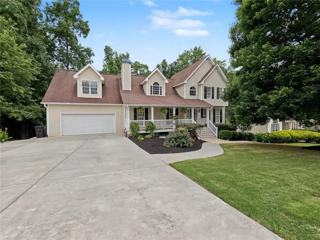 Traditional-style residence featuring light-colored siding, a prominent front porch with white railings, a two-car garage, dormer windows, and a concrete driveway