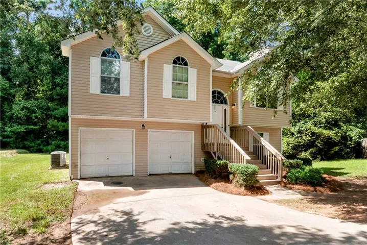 Two-car garage with white panel doors, elevated entry with wooden deck and stair access, tan vinyl siding exterior, gabled roofline with multiple window placements including arched transom details, and mature tree-lined landscaping surrounding the property