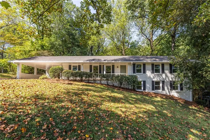 Split-level residence featuring a carport, white brick exterior, black window shutters, and a sloped lawn with mature trees