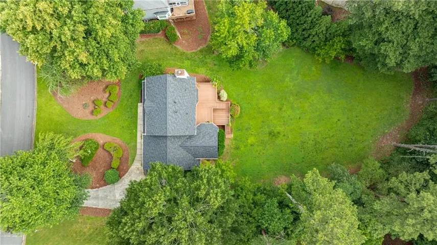 Expansive property featuring a dark shingle roof, a large wooden deck, a well-maintained lawn, mature perimeter trees, and a concrete driveway with landscaped beds