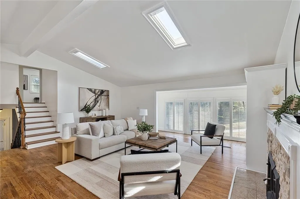 Living room with a stone fireplace, vaulted ceiling with beams, light wood finished floors, and a skylight