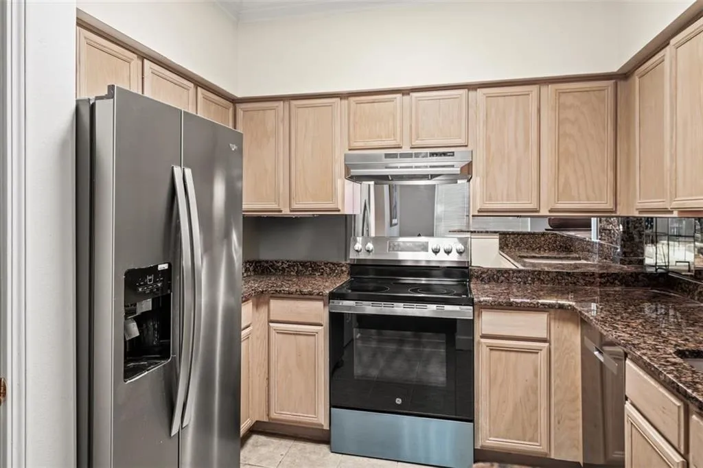 Kitchen with light brown cabinets, appliances with stainless steel finishes, light tile patterned floors, under cabinet range hood, and dark stone countertops