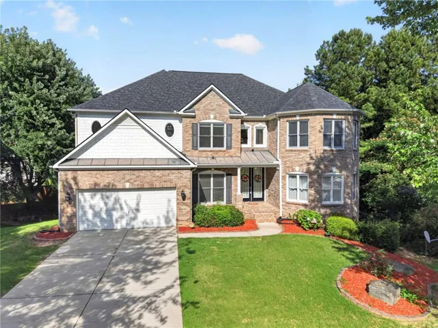 Two-story brick facade residence with a two-car garage, dark shingle roof, and a mature tree-lined perimeter