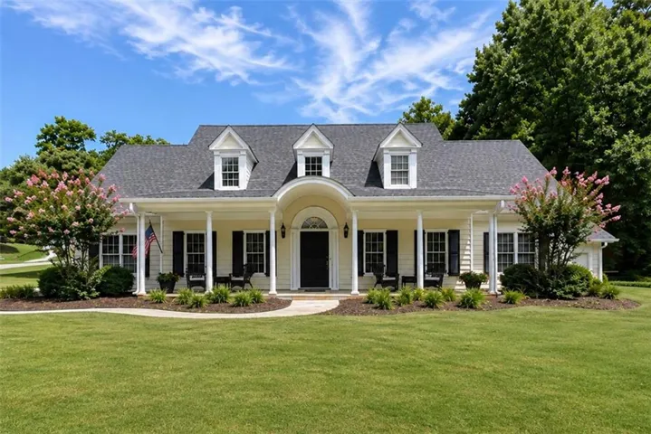 Manicured front lawn with a curved walkway, covered front porch, dormer windows, dark shutters, and mature landscaping