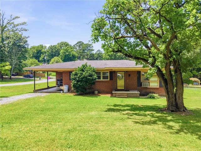 Brick ranch-style home featuring a covered carport, gravel driveway, and a mature tree providing shade to the front yard