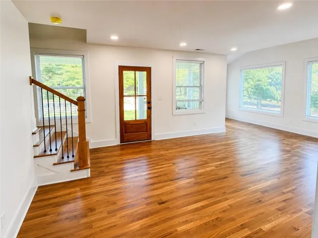 Spacious interior featuring wood-finish flooring, a wood staircase with black iron spindles, multiple windows with blinds, a paneled wood entry door, and recessed ceiling lighting