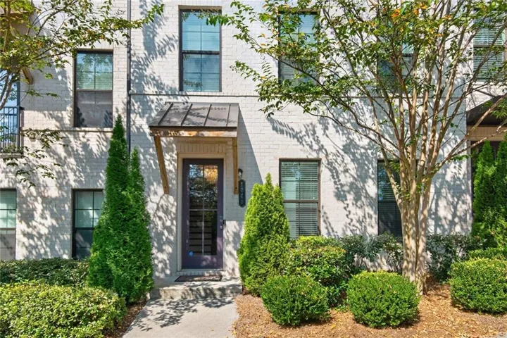 White brick exterior with multiple windows, architectural awning, dark paneled entry door, and mature landscaping