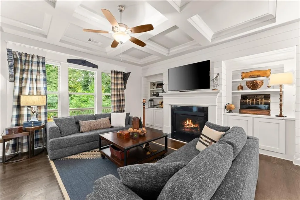 Living area featuring dark wood-type flooring, a glass covered fireplace, beam ceiling, a ceiling fan, and built in shelves