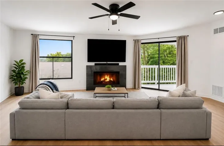 Spacious living area featuring wood-finish flooring, a contemporary fireplace with dark surround, a ceiling fan, and sliding glass doors opening to an outdoor balcony