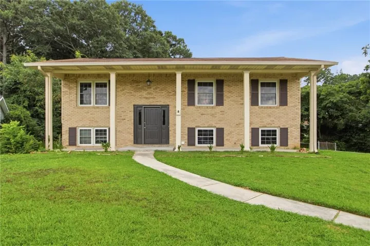 Brick facade home featuring a covered entryway with four support columns, dark paneled double entry doors, multiple windows with dark shutters, and a concrete pathway through a green lawn