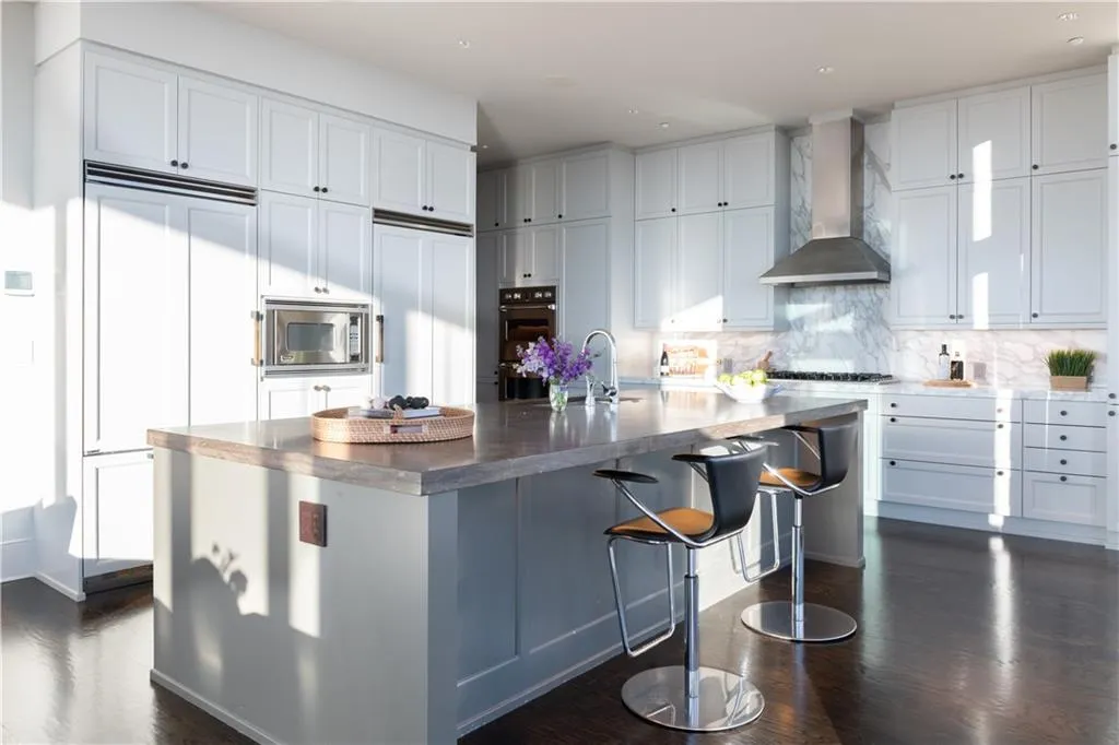 Kitchen featuring a spacious island, wall chimney exhaust hood, built in appliances, decorative backsplash, and white cabinets