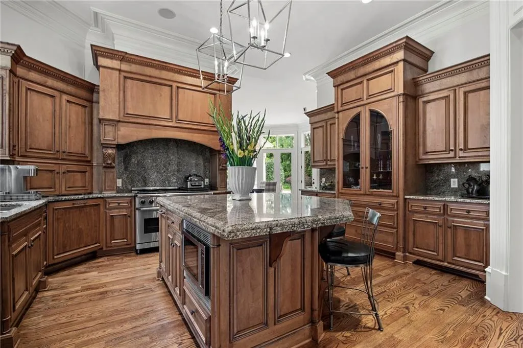 Gourmet kitchen featuring custom wood cabinetry with raised-panel doors, granite countertops, a large center island with seating, stainless steel appliances, dark stone backsplash, hardwood flooring, and a geometric pendant light fixture suspended above the island