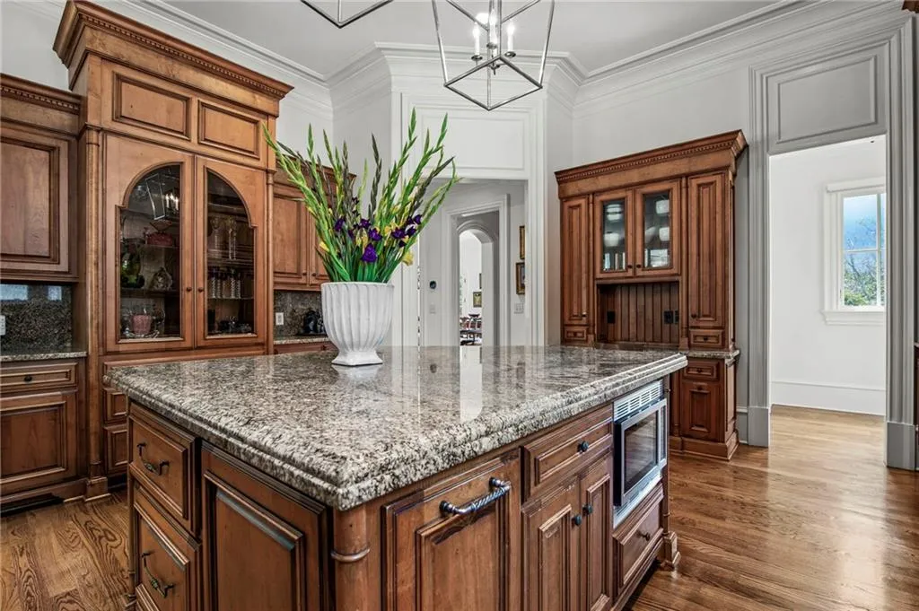 Kitchen island featuring granite countertop, built-in microwave, wood-finish flooring, and custom wood cabinetry with glass-front uppers