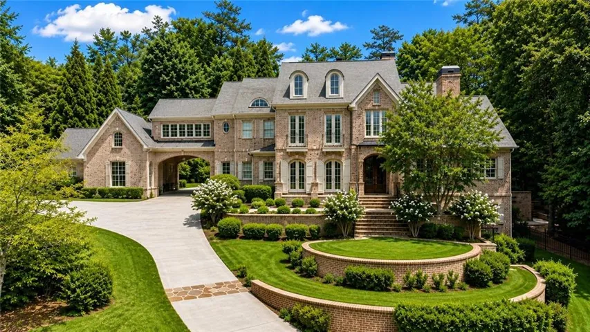 Brick exterior with multiple dormer windows, a porte-cochère, a winding concrete driveway, and tiered landscaping with a brick retaining wall