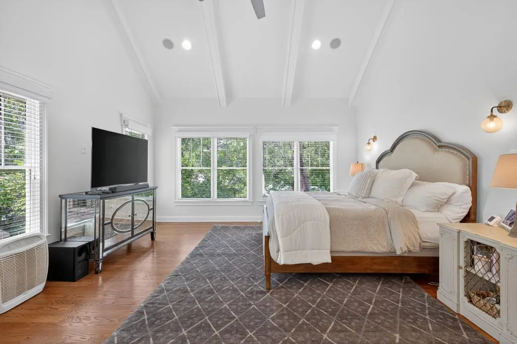 Vaulted ceiling with exposed beams, recessed lighting, wood-finish flooring, multiple windows, and wall-mounted light fixtures