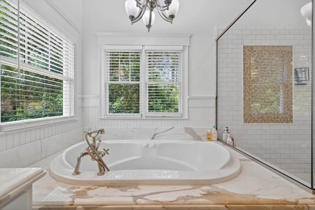 Luxurious primary bathroom featuring a curved soaking tub with wood-finish surround, subway tile walls, a frameless glass-enclosed shower, and abundant natural light from multiple windows overlooking landscaped grounds