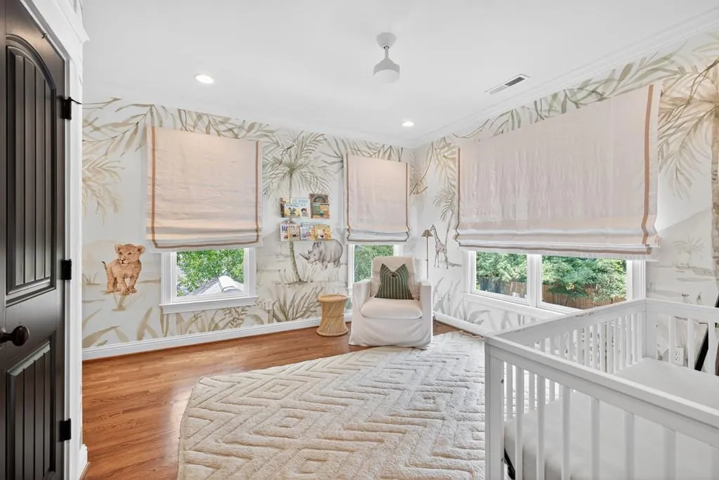 Bright nursery showcasing hardwood flooring, multiple windows with roman shade treatments, botanical wallpaper detailing, and white cabinetry
