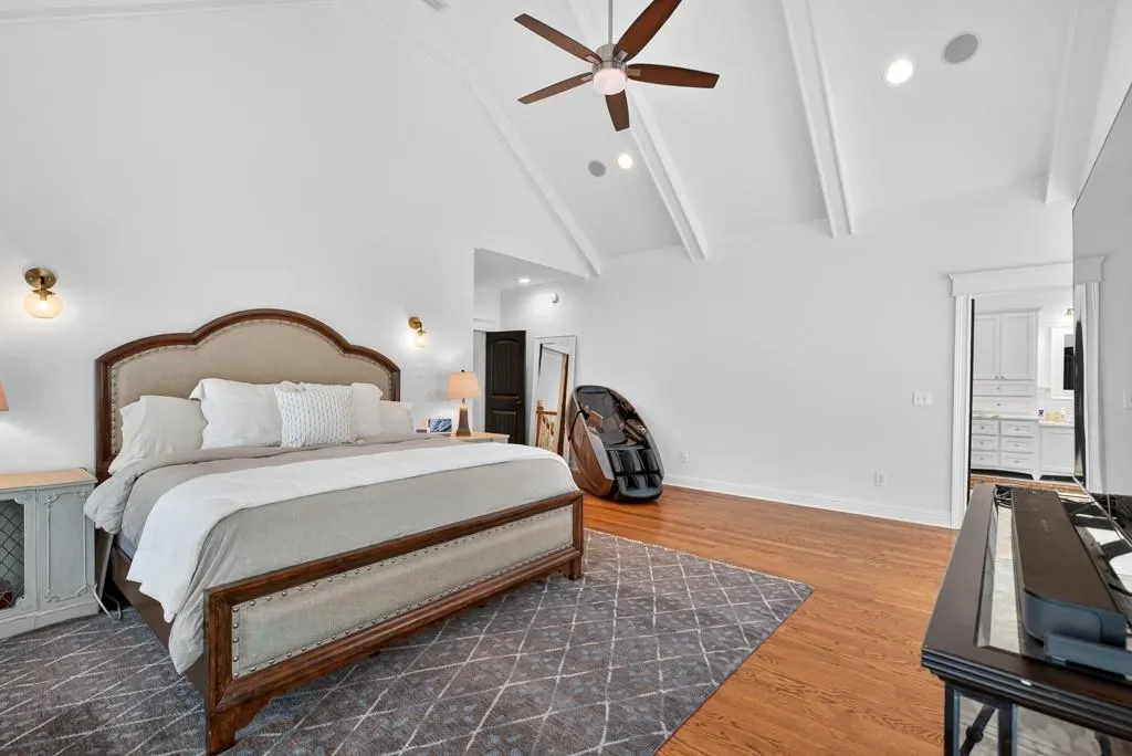 Spacious primary bedroom suite featuring vaulted ceilings with exposed beams, a wood-frame bed with upholstered headboard, hardwood flooring, recessed lighting, a ceiling fan with dark blades, and an en-suite bathroom entry